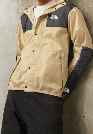 Mężczyzna w beżowo-czarnej kurtce North Face z logo na piersi i rękawie, w połączeniu z czarnymi spodniami, stoi przy betonowej ścianie.