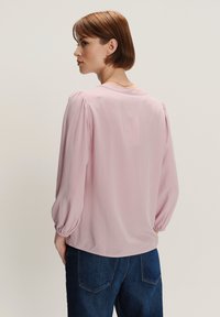 Donna con capelli corti castani di spalle, che indossa una blusa rosa chiaro a maniche lunghe e jeans blu scuro su uno sfondo semplice.