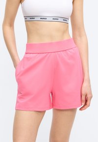 Γυναίκα που φοράει φωτεινά ροζ σορτς με ψηλή μέση και λευκό αθλητικό σουτιέν με ρέλι λογότυπου "HUGO", με το χέρι στην τσέπη, στέκεται μπροστά σε λευκό φόντο.