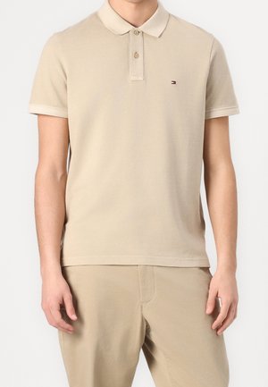 Homme portant un polo à manches courtes beige avec un petit logo sur la poitrine gauche et un pantalon beige assorti, debout devant un fond uni.