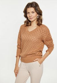 Sweter w serek w jasnobrązowym kolorze z diamentowym wzorem i ściągaczem na dole. Zestawiony z beżowymi spodniami z bocznym detalem kieszeni.