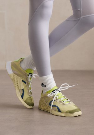 Osoba nosząca jasnoszare legginsy, białe skarpety do kostek oraz żółto-zielone sportowe buty z białymi sznurówkami, stojąca na beżowej podłodze.