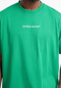 Tamsaus odos atspalvio žmogus vilki ryškiniai žalią marškinėlį su baltais užrašu "GUESS JEANS" centriniame krūtinės srityje.