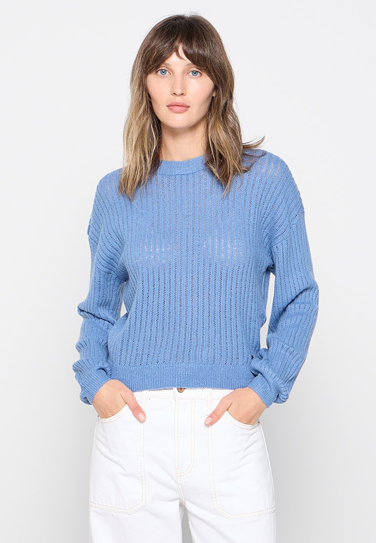 Lichtblauwe gebreide trui met een ribbelstructuur, ronde halslijn en lange mouwen. Gekozen met witte hoge taille broek.