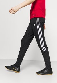 Black athletic pants with white three-stripe design, paired with black shoes featuring textured surface and yellow lettering, worn with a red shirt.