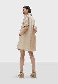 Abito di cotone beige con maniche corte e colletto, caratterizzato da una vestibilità ampia e tasche. Indossato con sandali a blocco con tacco dorati.