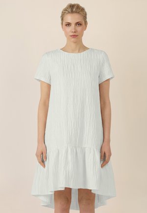 Blondynka ubrana w jasnoniebieską, teksturowaną sukienkę z krótkim rękawem i asymetrycznym dołem, na jednolitym beżowym tle.