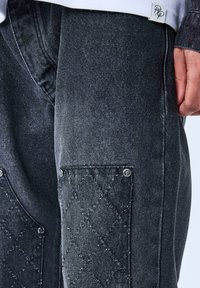 Pantalones de mezclilla en gris oscuro con un bolsillo acolchado texturizado. Presenta herrajes plateados y un ligero brillo en la tela.