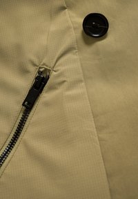 Bézs, texturált anyag átlós rácsmintával, fekete cipzárral és gombbal. Tiszta varrások és minimalista design elemek.