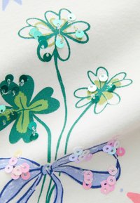 Kwiatowy wzór na tkaninie z zielonymi i zielononiebieskimi kwiatami, ozdobiony cekinami. Jasnoniebieska wstążka z różowymi cekinami u podstawy.