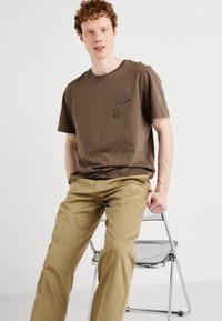 Short brown t-shirt with a round neckline and two small skull illustrations on the left chest, paired with light beige trousers.