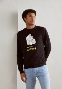 Sudadera negra con un gráfico de dibujos animados de un personaje sentado en una silla, con texto amarillo que dice "Los Simpson". Ajuste casual, puños acanalados.