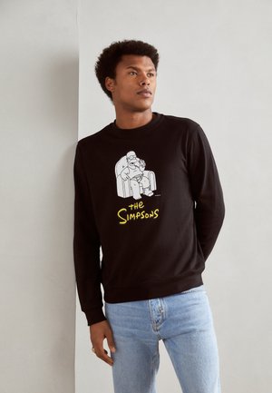 Sudadera negra con un gráfico de dibujos animados de un personaje sentado en una silla, con texto amarillo que dice "Los Simpson". Ajuste casual, puños acanalados.