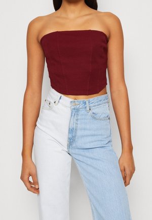 Kobieta w bordowym, bezramieniowym crop topie, zestawionym z wysokim stanem, półbiałymi, półjasnoniebieskimi jeansami na neutralnym tle.