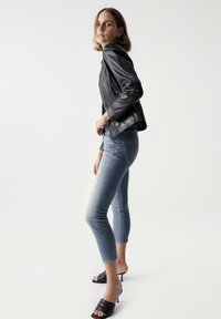 Schwarze Lederjacke mit Reißverschlusstaschen, hellblaue, verkürzte Jeans mit used-Look und schwarze, hochhackige Mules.