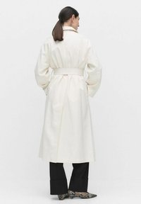 Biały płaszcz trench z paskiem w talii, obniżonymi szwami na ramionach i długą sylwetką. Noszony na czarnych spodniach, prezentujący luźny styl.