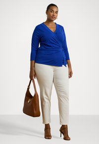 Blue wrap top with three-quarter sleeves, draped front detail; paired with cream fitted trousers and brown handbag. Smooth fabric texture.