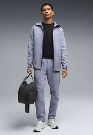 Jonge man in een lichtgrijs trainingspak en zwart shirt met een zwarte sporttas, staand tegen een grijze verloopachtergrond.