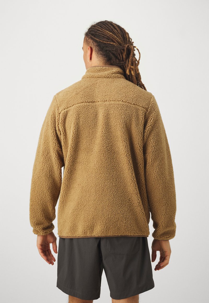 Giacca di pile marrone con un materiale testurizzato e peloso e colletto alto. Il modello indossa pantaloni corti scuri e ha i capelli lunghi raccolti.