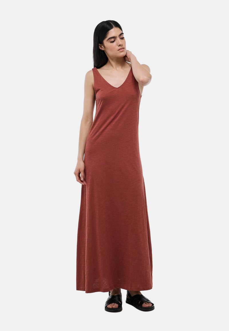Robe maxi de couleur rouille, en tissu doux. Elle présente un décolleté en V, sans manches et une coupe ample, atteignant les chevilles. Sandales noires portées.