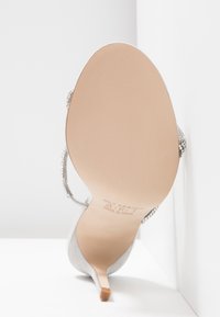 Sandale à talon haut grise avec talon fin, dessus lisse et lanières ornées de strass. Comprend une semelle beige avec le logo sur le dessous.