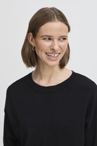 Pullover nero con collo rotondo e dettagli a costine, caratterizzato da una vestibilità rilassata. La modella ha capelli corti e orecchini a cerchio dorati.