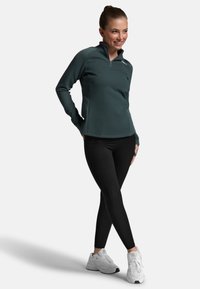 Mørkegrøn zip-up træningstrøje med teksturerede ærmer, parret med sorte leggings og hvide sneakers. Har et tætsiddende design og høj krave.