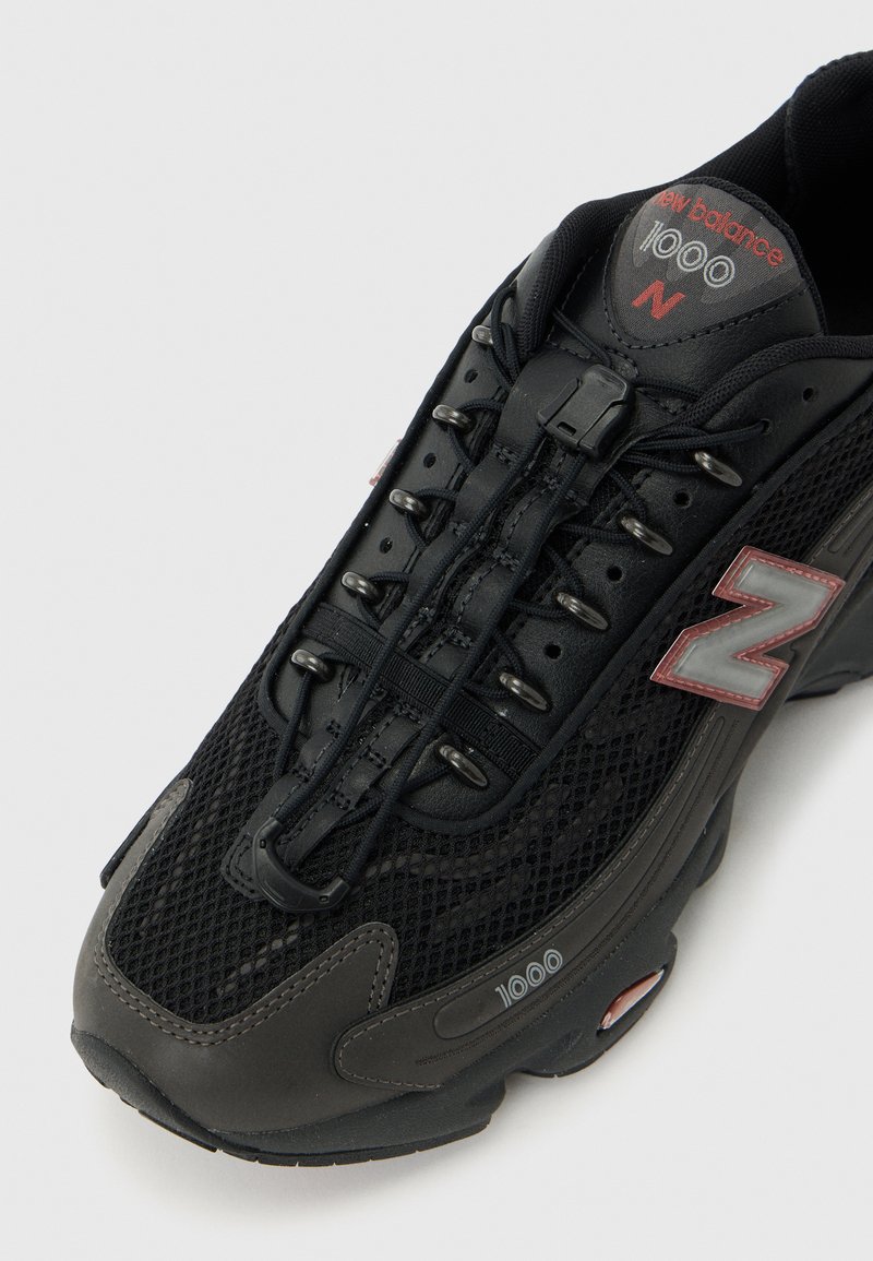 Zapatilla New Balance 1000 negra con parte superior de malla, logotipo reflectante "N", sistema de cierre rápido y detalles grises en la puntera y la suela.