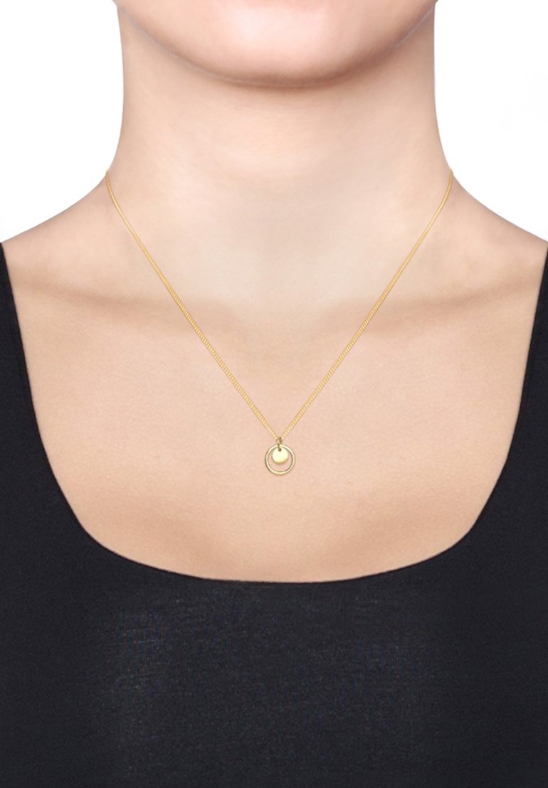 Collier en or avec un pendentif circulaire comportant un cercle plus petit à l'intérieur. Chaîne fine, design simple, porté au-dessus d'un haut noir sur un fond uni.