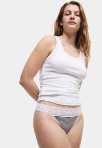 Weißes geripptes Tanktop mit Rundhalsausschnitt, kombiniert mit einem gestreiften Tanga mit einem weißen Spitzenbund. Das Model posiert mit den Händen in den Hüften.