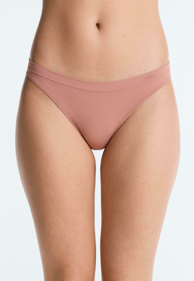 Vrouw draagt lichtbruine naadloze bikini-onderkleding, getoond van midden torso tot bovenbenen tegen een effen achtergrond.