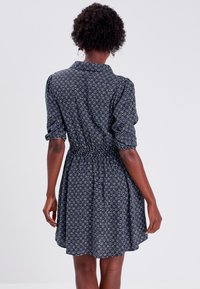 Femme aux cheveux bouclés portant une robe bleu marine avec des motifs géométriques blancs, se tenant de dos contre un fond uni.