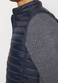 Navy blue padded vest with high collar and zip closure, paired with a textured gray sweater. Vest features horizontal stitching and orange zipper pull.