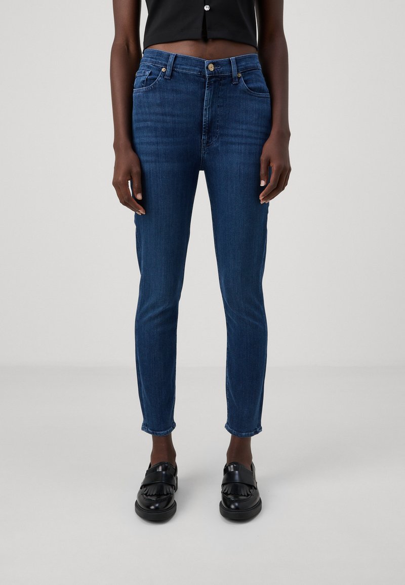 Jean en denim ajusté de couleur bleu foncé, avec cinq poches, une fermeture à bouton et un léger décoloration à l'avant, associés à des mocassins noirs.