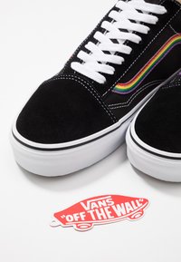 Svarte semskede sneakers med hvite snører, med en flerfarget sidekode. Inkluderer en rød Vans "Off the Wall" logoetikett nedenfor.