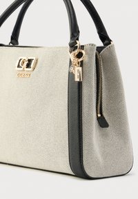 Bézs kézitáska fekete részletekkel, texturált anyagból, arany színű fémrészekkel, cipzárral záródik, és logó-emblémával díszített. Tartalmaz egy díszítő medált is.