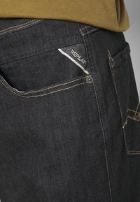 Vaqueros de denim oscuro con un bolsillo trasero que tiene una etiqueta blanca. Costuras en contraste y un acento de botón en la cinturilla. Textura suave.