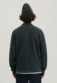 Donkergroene gebreide trui met een gestructureerd patroon, ronde hals en ribgebreide manchetten. Een bijpassende beanie wordt gedragen.