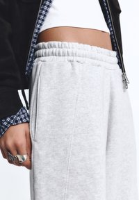 Persoon in lichtgrijze sweatpants, witte cropped top, zwarte jas, en een zichtbaar geruite shirtmouw, met ringen aan de linkerhand.