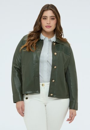 Donna con capelli castani ondulati che indossa una giacca di pelle verde scuro con bottoni sopra una camicia bianca a righe e pantaloni bianchi con bottoni decorativi.