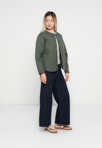 Femme portant une veste matelassée verte, une chemise gris clair, un jean large foncé et des baskets beiges à imprimé léopard, se tenant devant un fond blanc.