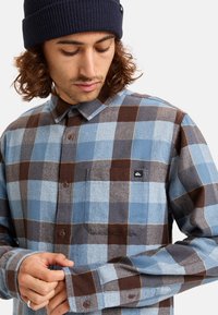 Camicia in flanella a quadri blu e marrone con design a bottoni, dotata di una tasca a toppa e di un'etichetta con logo sul petto. Tessuto testurizzato.