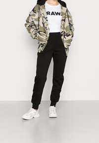 Maskovací bunda s černou kapucí, bílý tričko s textem "RAW." a černé joggingové kalhoty. Doplněno o bílé tenisky s texturovanou podrážkou.