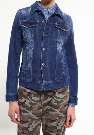 Personne portant une veste en jean bleue avec fermeture à boutons et un pantalon à motif camouflage, debout devant un fond blanc.