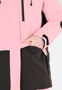 Rosa wasserdichte Jacke mit hohem Kragen, schwarzem unteren Teil, Reißverschlusstasche und elastischen Bündchen. Strukturiertem Material mit glatter Oberfläche.