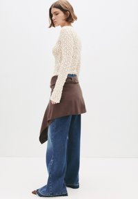 Vrouw met kort bruin haar poseert zijwaarts in een crèmekleurige kanten top met lange mouwen, een bruin asymmetrisch rokje over wijdvallende blauwe jeans.