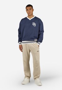 Pullover de algodão azul-marinho com decote em V branco e logotipo, combinado com calças beige com nervuras e ténis brancos com acabamentos texturizados.