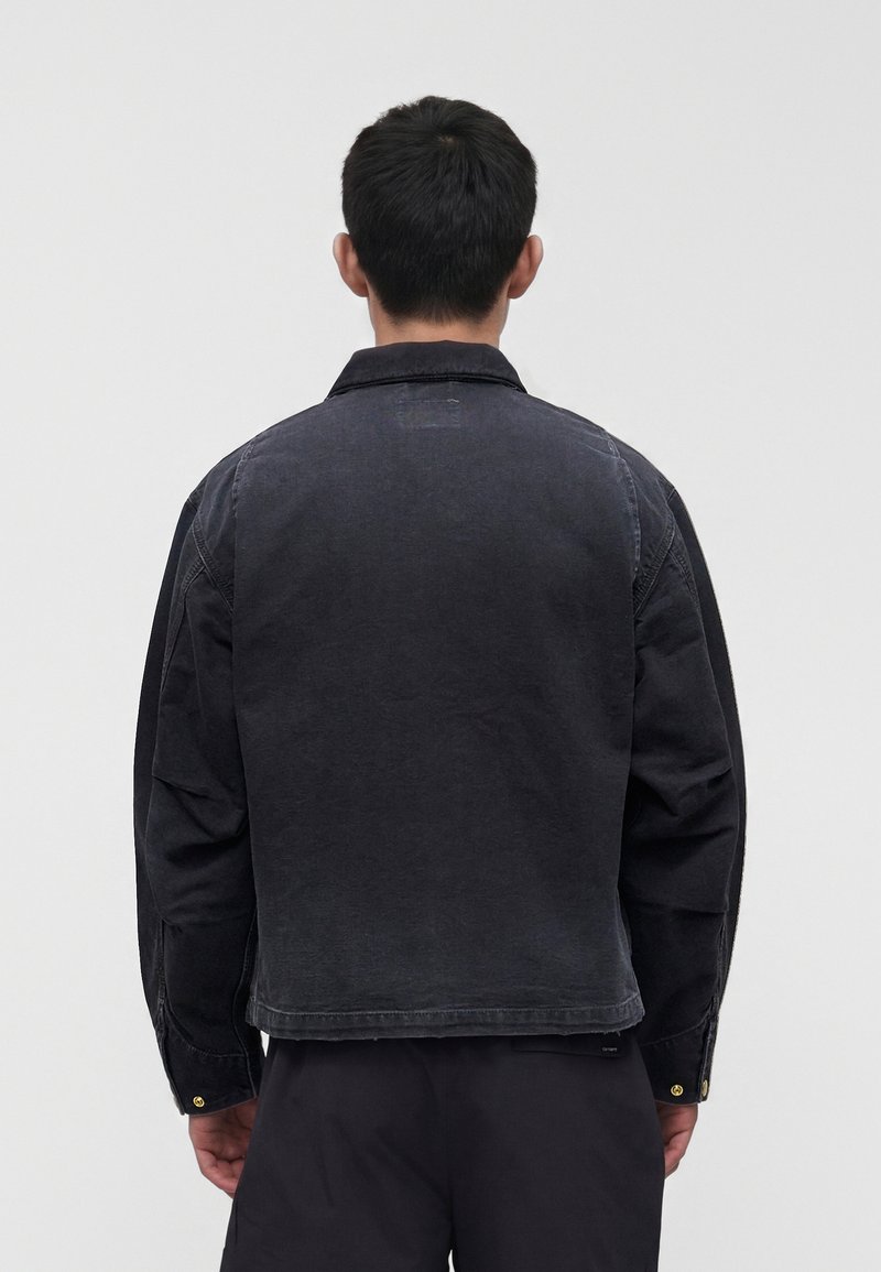 Mann mit kurzen schwarzen Haaren steht mit dem Rücken zur Kamera, trägt eine schwarze Jeansjacke und schwarze Hose vor einem einfarbigen hellen Hintergrund.