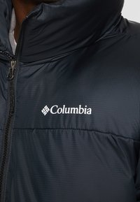 Czarna puchowa kurtka Columbia z wysokim kołnierzem, zamkiem błyskawicznym i białym logo na klatce piersiowej. Wykonana z gładkiego, teksturowanego materiału.