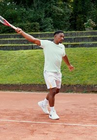 Sportlane kergrohelises särgis ja valgetes lühikeses pükstes, millel on oranžid aktsendid, mängib tennist savi väljakul, hoides racketit.